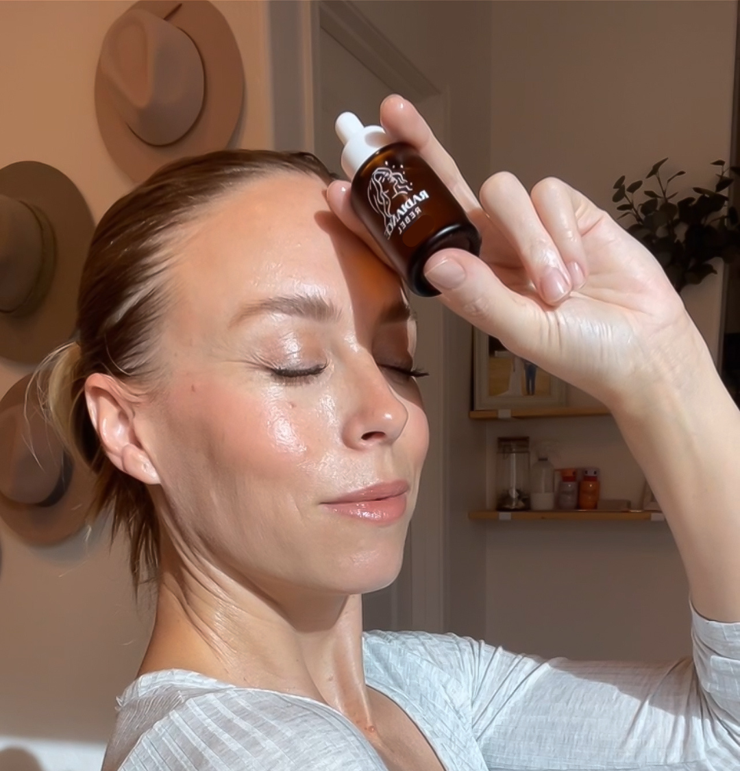 Woman applying a dropper of a brown liquid to her face in a home setting.