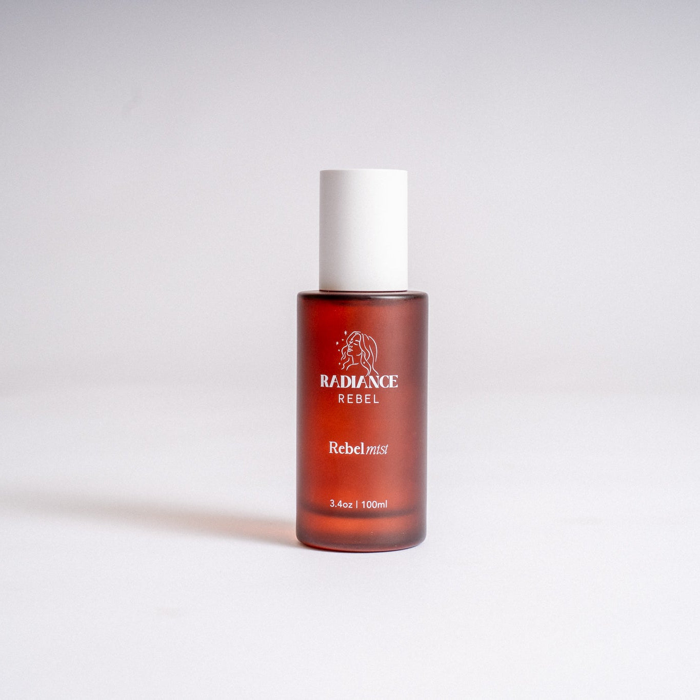 A 2 fl oz amber glass bottle of RebelMist by Radiance Rebel with a white cap, standing upright on a light gray background.