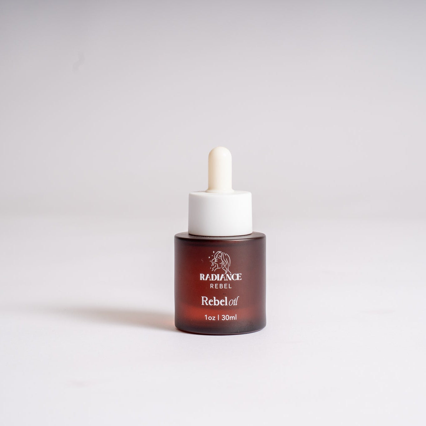 A small amber glass dropper bottle with a white rubber pipette top and a white collar, labeled 'RADIANCE REBEL' and 'Rebeloil', centered on a light gray studio background.
